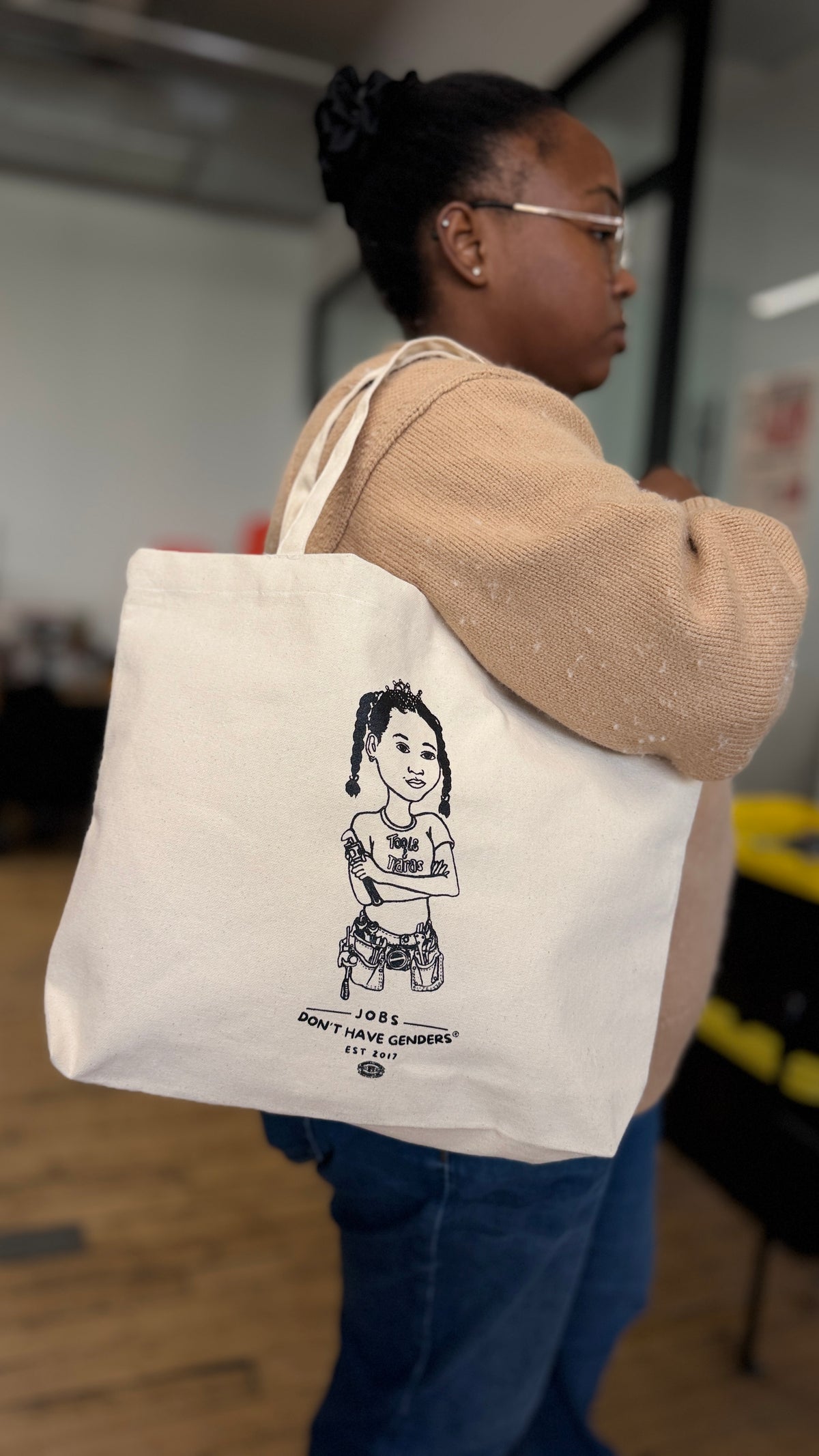 Person holding the Jobs Don't Have Genders® off-white canvas tote bag from Tools & Tiaras® to show its size